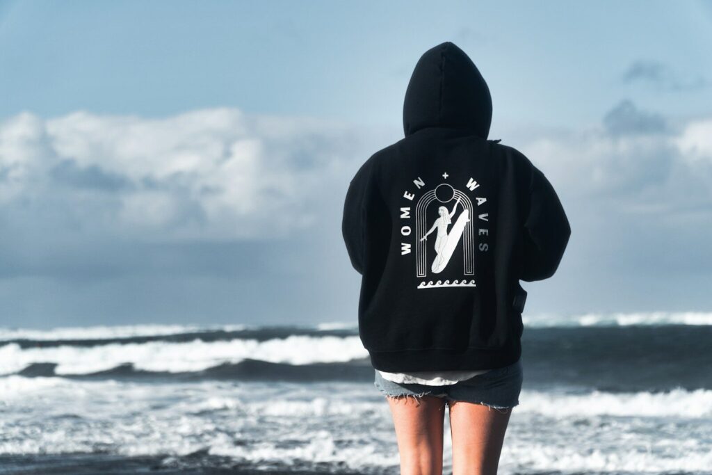 Person in hoodie facing ocean waves on beach
